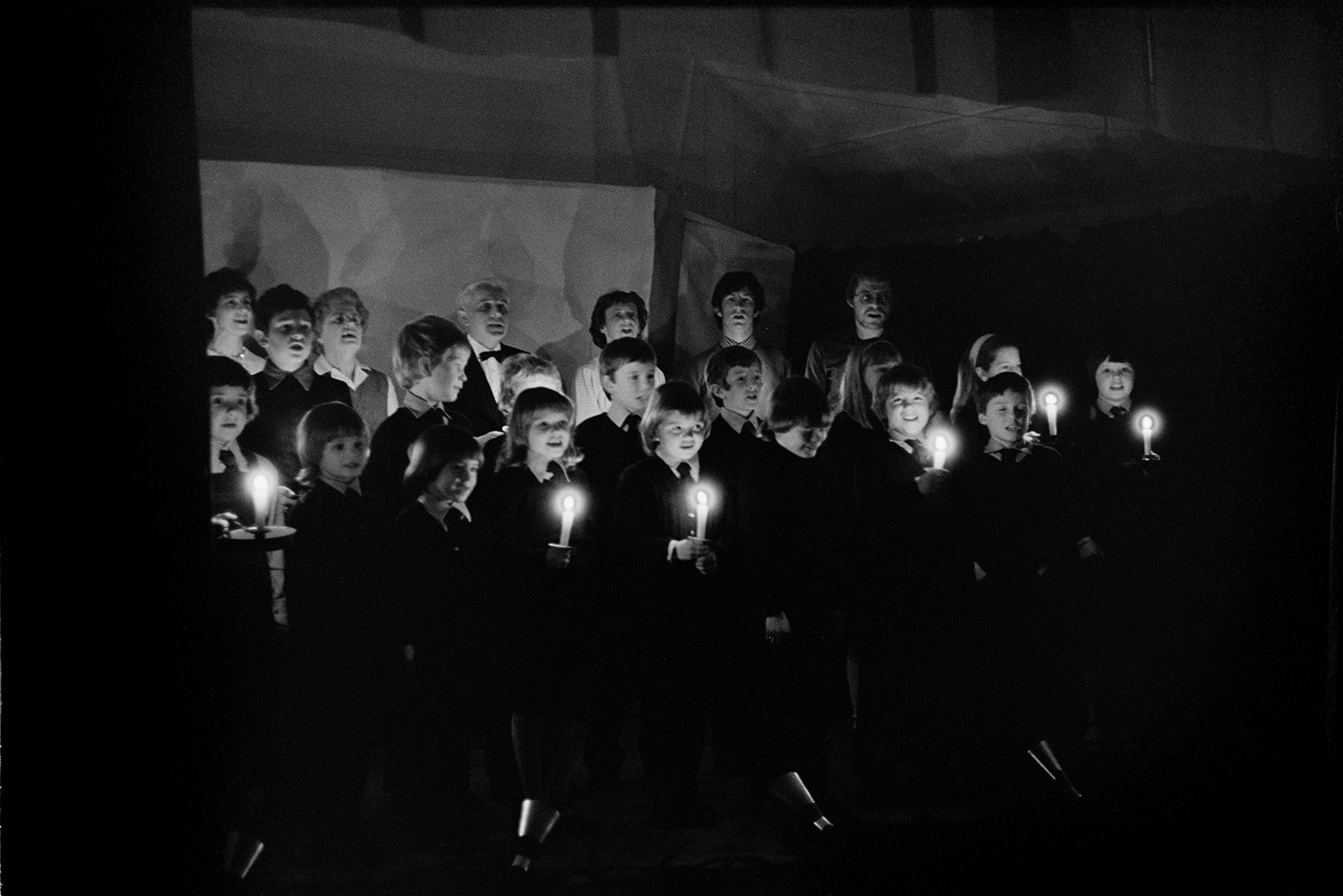 Christmas entertainment in village hall, singing and dance, short plays.
[Children and adults singing on a stage at High Bickington village hall at an evening of Christmas entertainment. Some of the children are holding lit candles.]