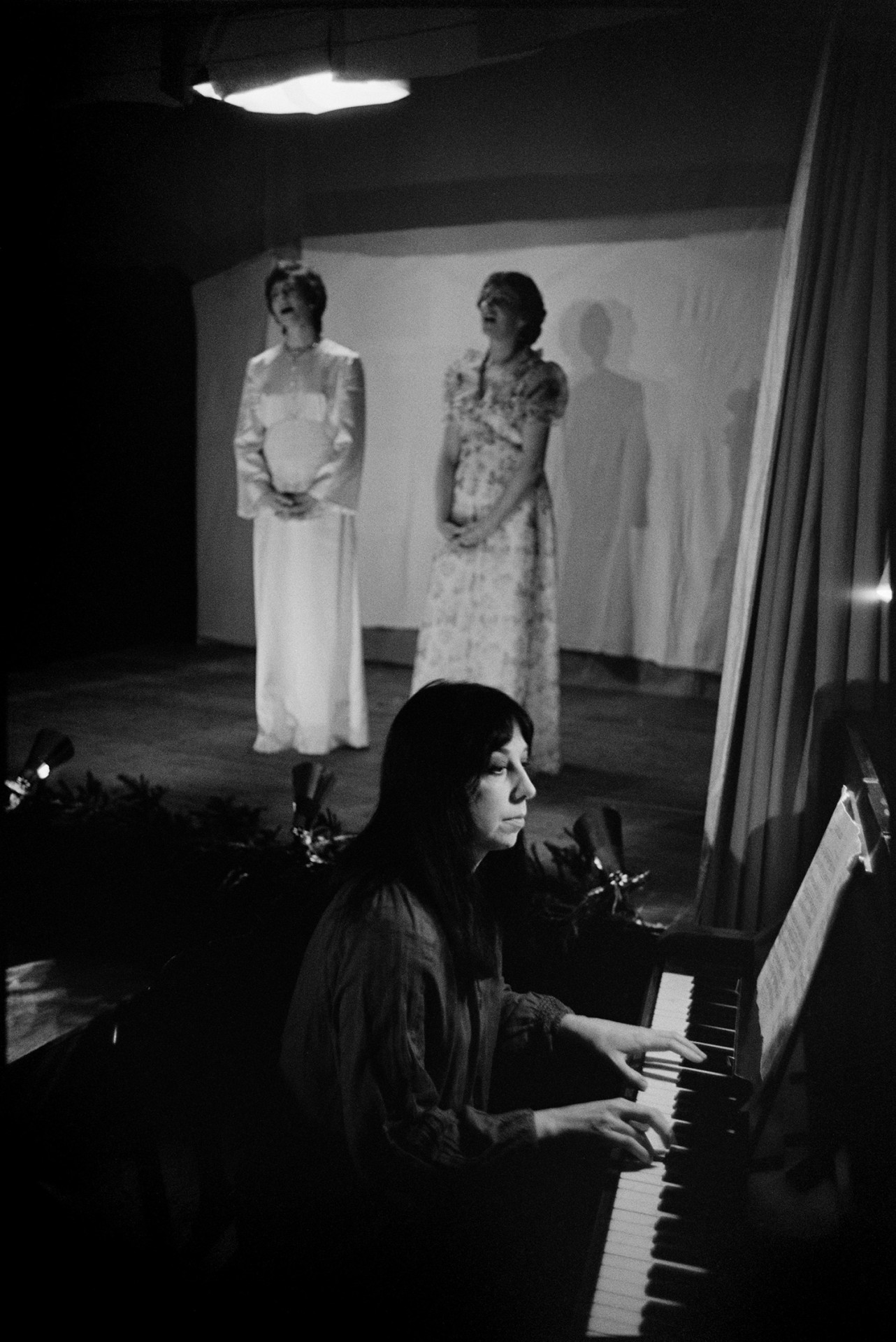Christmas entertainment in village hall, singing and dance, short plays.
[Two women singing on stage at High Bickington village hall at an evening of Christmas entertainment. A woman in the foreground is accompanying them of the piano.]
