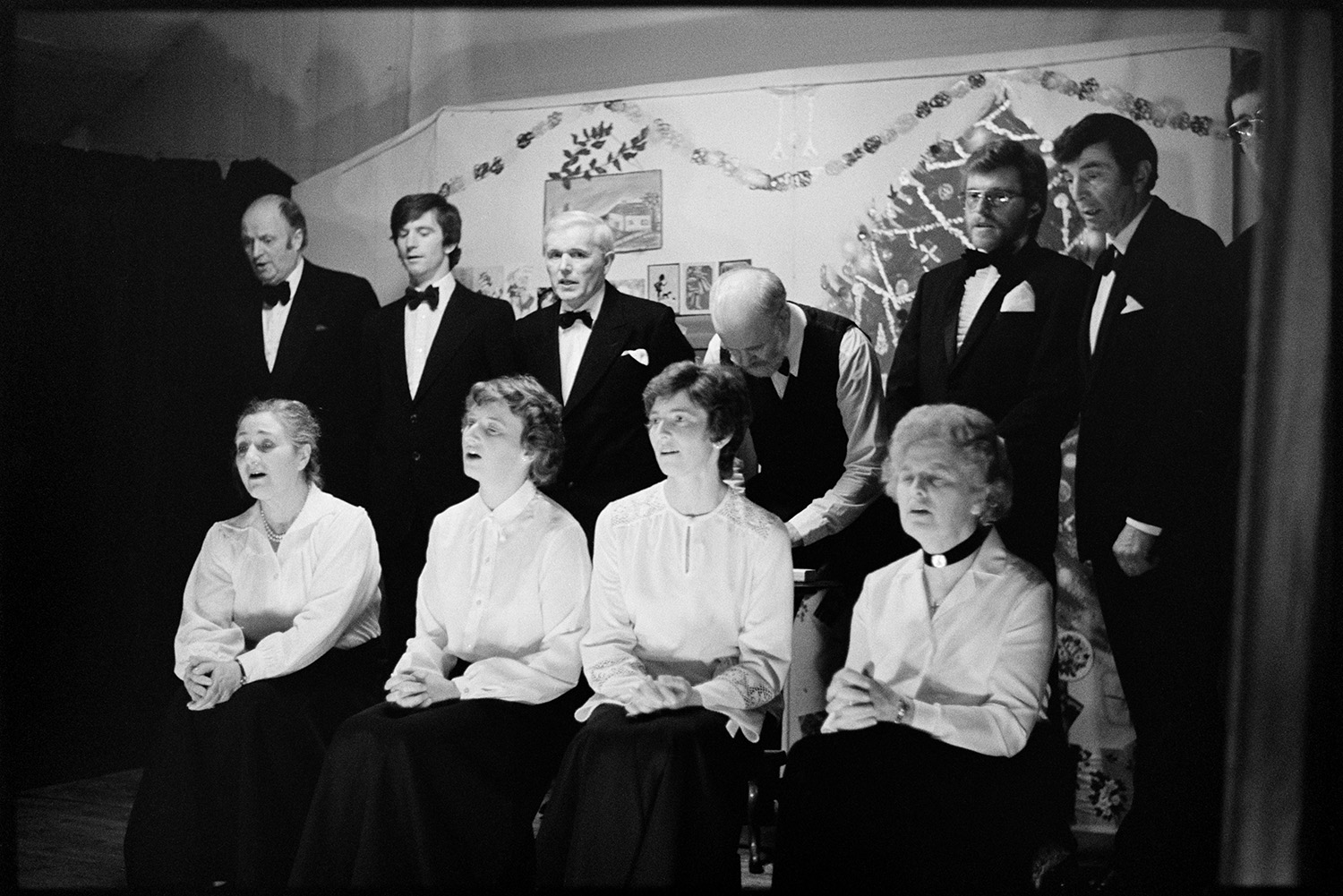 Christmas entertainment in village hall, choir singing to pianist.
[A choir of women and men singing on stage at High Bickington village hall at an evening of Christmas entertainment. The women are all wearing white shirts and the men are wearing bow ties.]