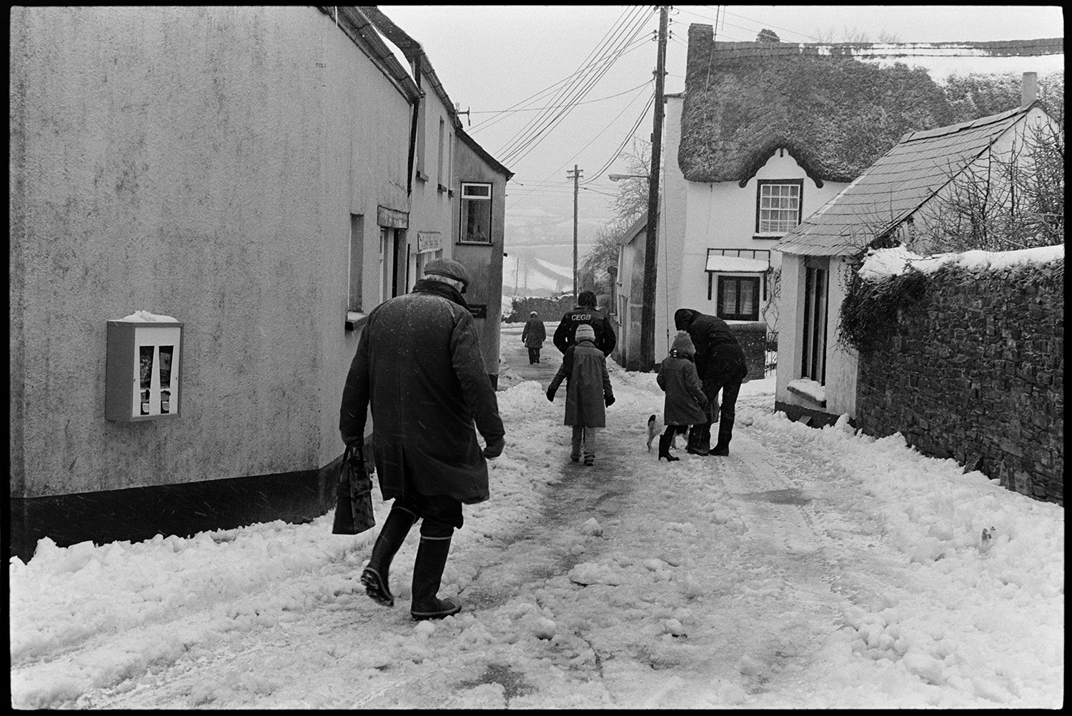Snow, street scene, shoppers. 
[People, including children, walking along Church Street in Dolton which is covered with snow. They are passing houses and a thatched cottage.]