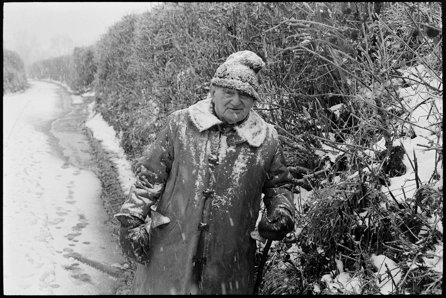 Snow, farmer and woman walking up lane, covered in snow. 
[Archie Parkhouse walking along West lane, Dolton which is covered in snow. He is wearing a woolly hat, a coat and gloves which have snow on them. Snow is falling.]