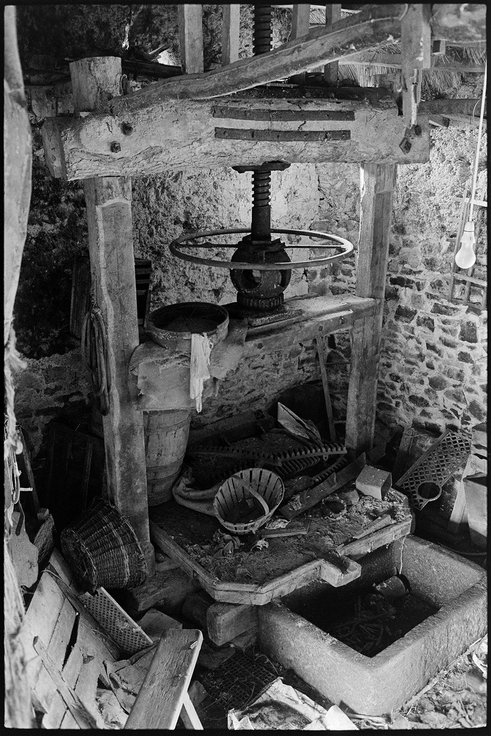 Old cider press, disused.
[An old, disused cider press in a cluttered cob and stone barn at West Chapple Farm, near Winkleigh. Various baskets are littered around the press.]