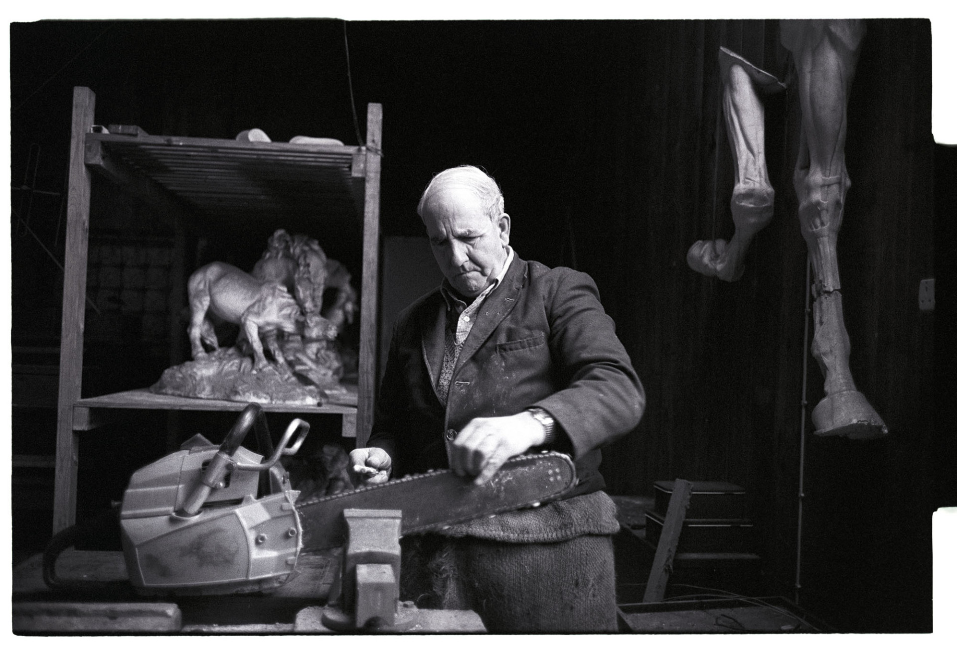 Man sharpening chain saw in workshop, formerly sculptors studio, with sculpture. 
[Horace Baker sharpening his chainsaw in his workshop at Halsdon House, Dolton. The workshop used to be a sculptors studio and two sculptures of horses are in the background.]