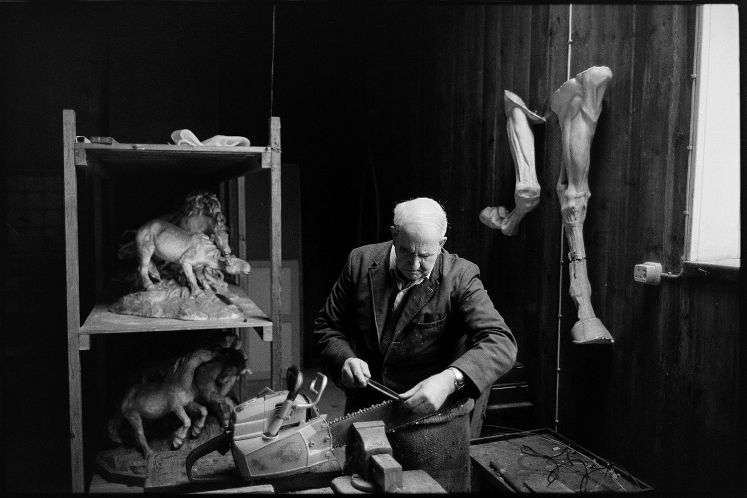 Man sharpening chainsaw in workshop, formerly sculptor's studio, with sculpture.
[Horace Baker sharpening a chainsaw, secured in a vice, in a workshop which was formerly a sculptor's studio, at Halsdon House near Dolton. Pieces of sculpture are displayed, including horses.]
