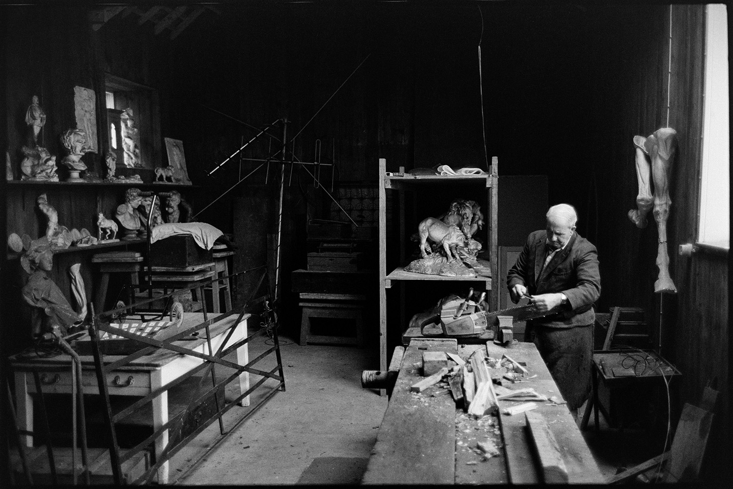Man sharpening chainsaw in workshop, formerly sculptor's studio, with sculpture.
[Horace Baker sharpening a chainsaw, secured in a vice, in a workshop which was formerly a sculptor's studio, at Halsdon House near Dolton. Pieces of sculpture are displayed, including horses and busts, along with a metal gate.]