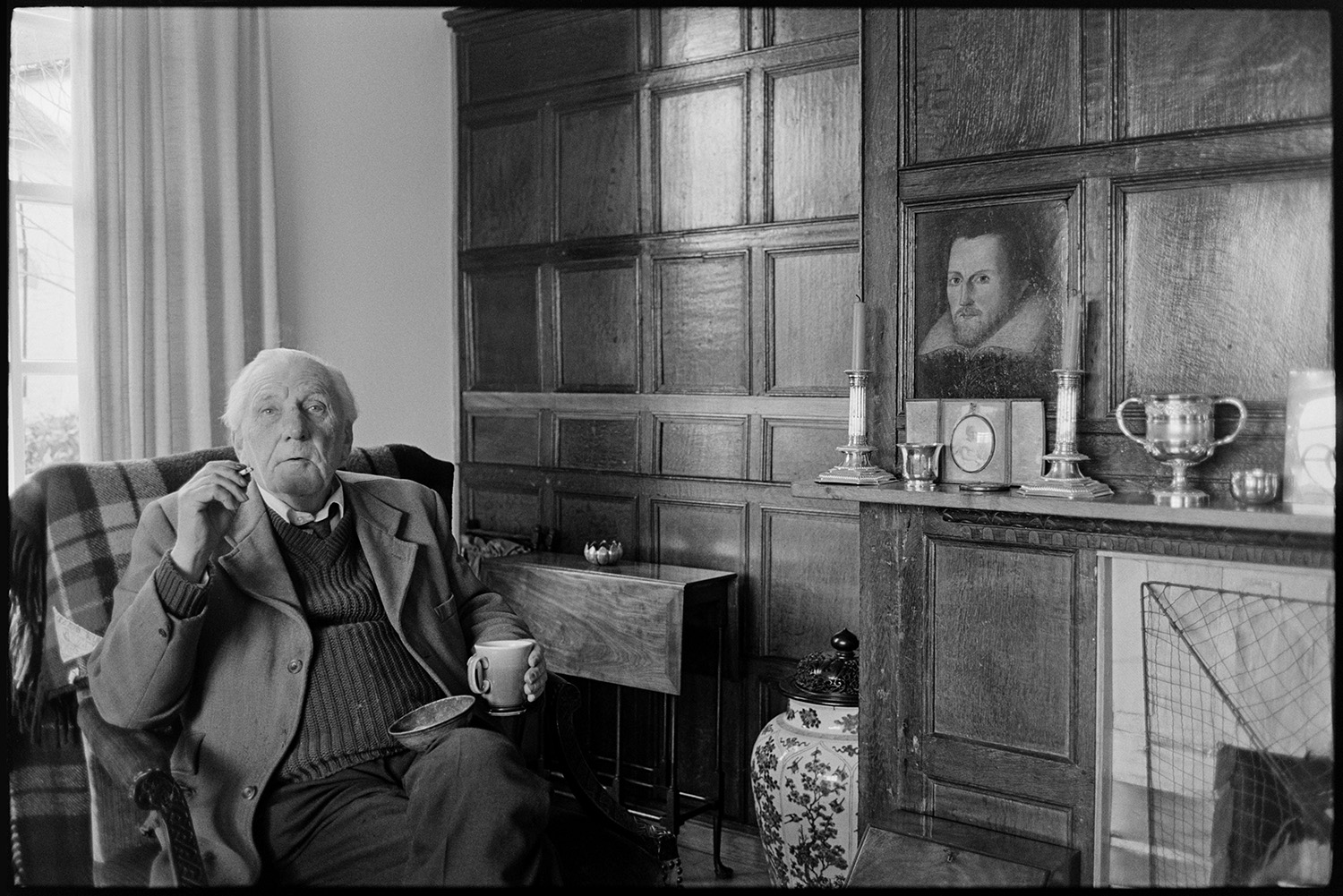 Man and woman seated, furniture, panelled screen. <br />
[Michael Davis, a retired solicitor, sitting in a chair by a fireplace at his house in Dolton. He is smoking a cigarette and holding a mug. The room has wooden panelling, and one of the panels contains a portrait of a man. Candlesticks and silverware are on display on the mantelpiece.]