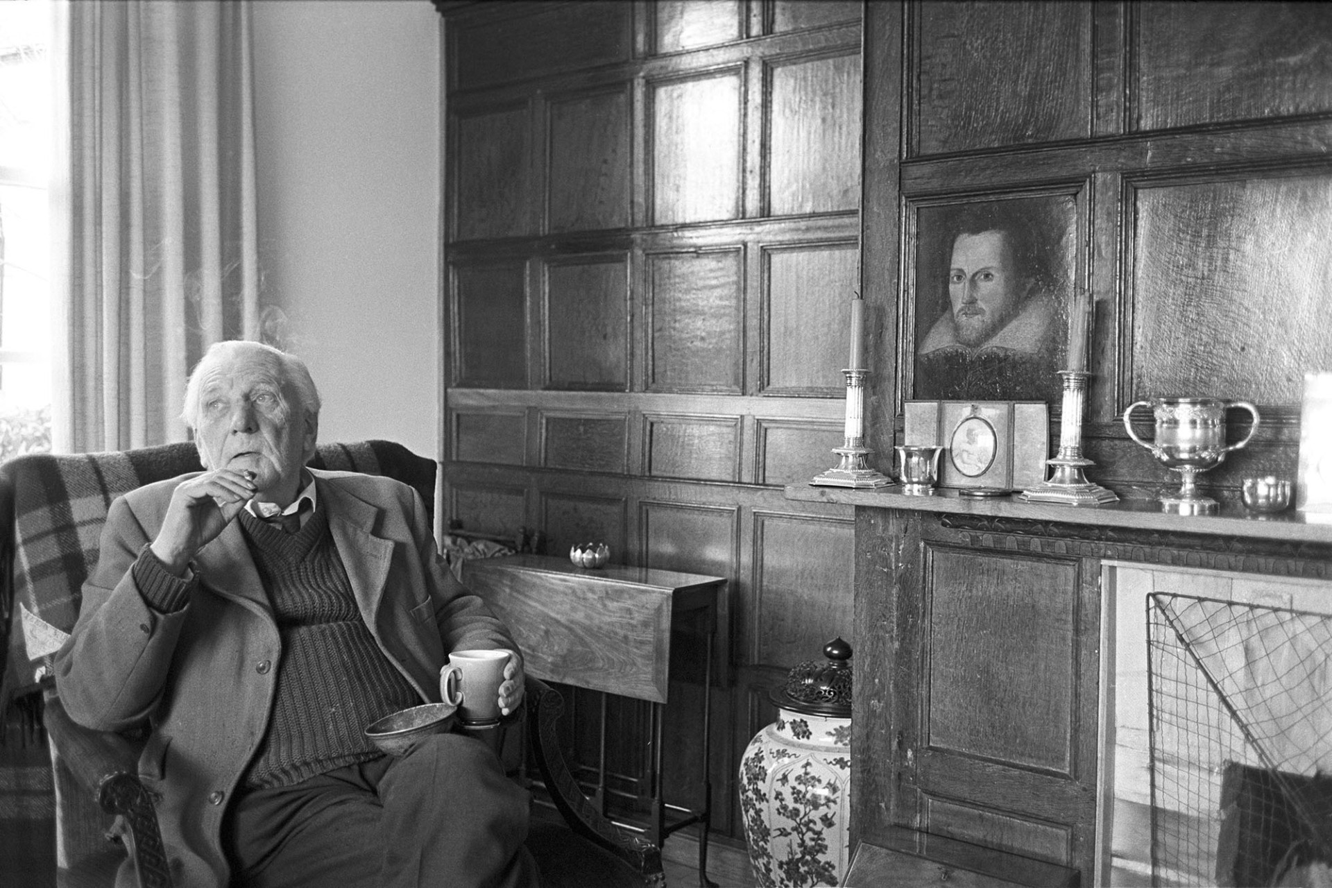 Man seated, smoking, furniture panelled screen with painting. 
[Michael Davis, a retired solicitor, sitting in a chair by a fireplace at his house in Dolton. He is smoking a cigarette and holding a mug. The room has wooden panelling, and one of the panels contains a portrait of a man. Candlesticks and silverware are on display on the mantelpiece.]