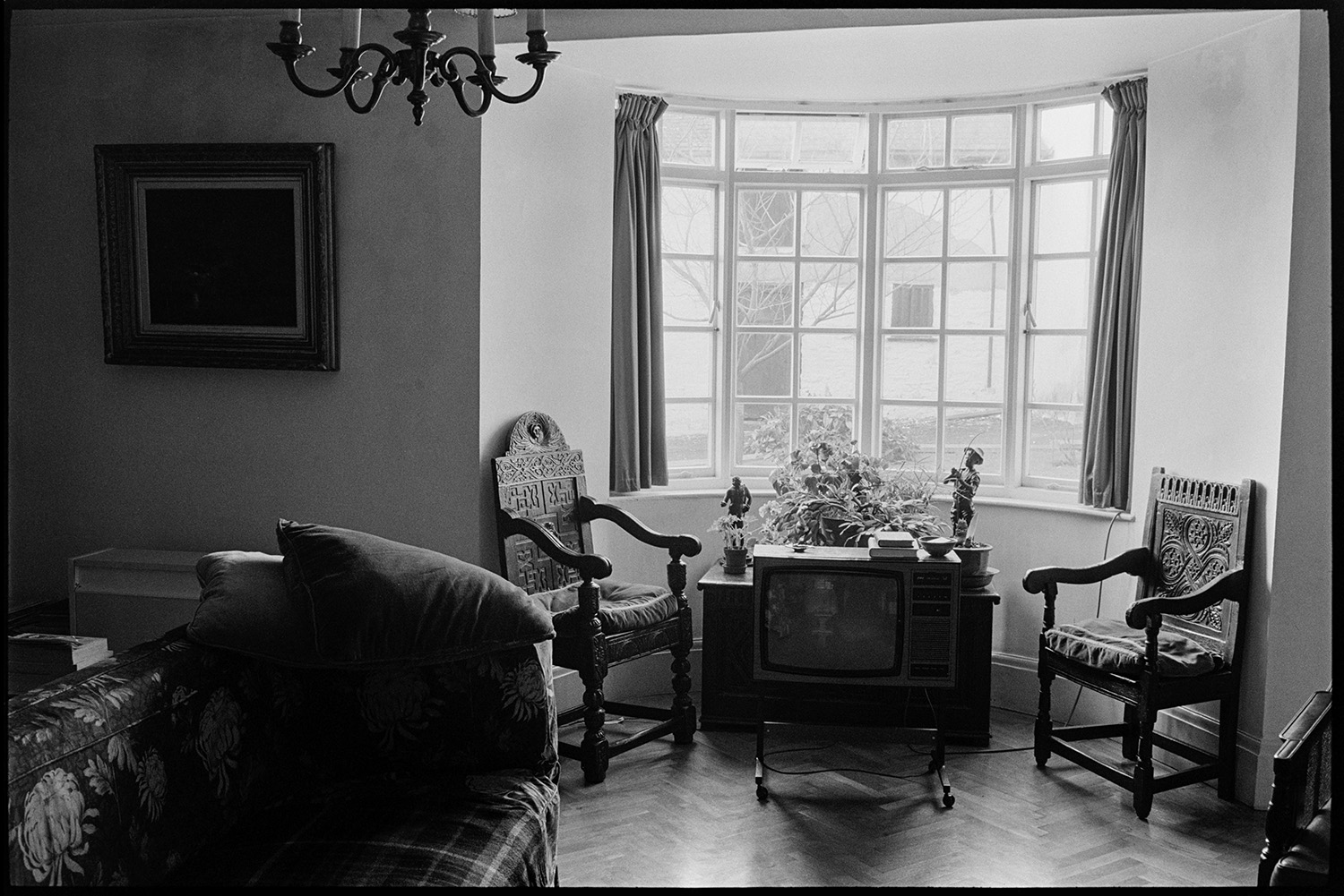 Man seated, smoking, furniture, china and paintings.
[Interior of a room in a house in Dolton, belonging to Michael Davis, showing a painting, two carved wooden chairs, a sofa and a television set, in front of a bay window.]