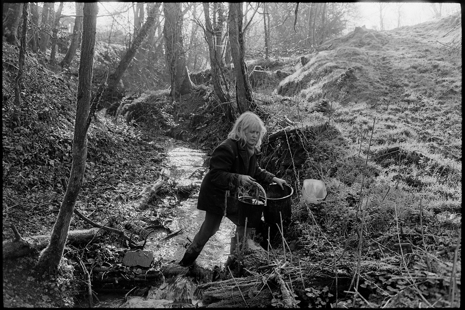 Woman farmer milking goat and fetching water from stream, old car in mud.
[Jo Curzon collecting water with plastic buckets, from a stream in a wooded valley at Millhams, Dolton.]