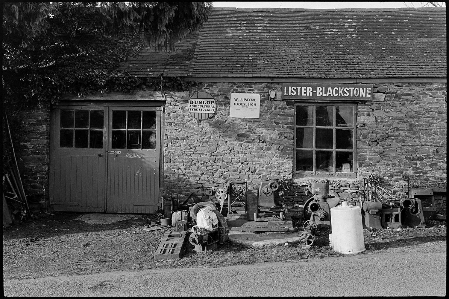 Front of engineer's workshop. 
[The front of W J Payne, Engineer's workshop at Iddesleigh. Various items and parts of machinery can be seen outside the workshop.]