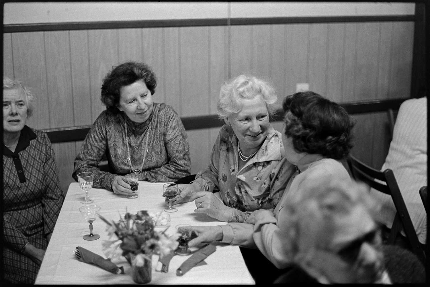 Women seated at W I dinner in village hall chatting. 
[Four women sat at a table talking and drinking at a Women's Institute dinner in Dolton Village Hall.]