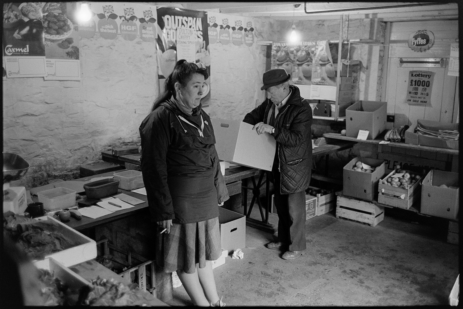 Woman serving in greengrocer's shop, waiting for customers. 
[Susan Jury running a greengrocer's shop in Dolton. She is smoking a cigarette. Various boxes of fruit and vegetables are displayed around the shop. Posters are pinned to one of the shop walls and a man is looking in one of the boxes.]