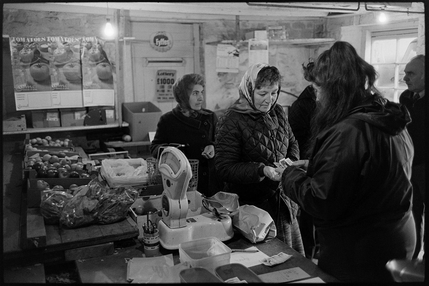 Woman serving in greengrocer's shop, waiting for customers. 
[Susan Jury handing a customer her change in a greengrocer's shop in Dolton. Other customers are queuing up to buy goods. Various boxes of fruit and vegetables are displayed around the shop, as well as posters advertising tomatoes.  A set of weighing scales can be seen on the counter.]