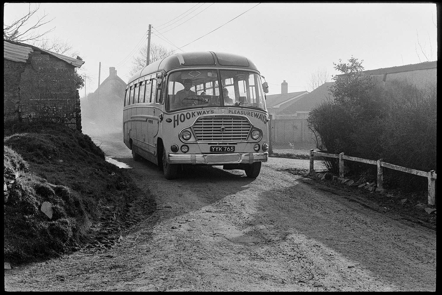 School bus arriving in village children waiting, morning. 
[A Hookways school bus driving along a road in Upcott, Dolton to pick up children  and take them to school.]