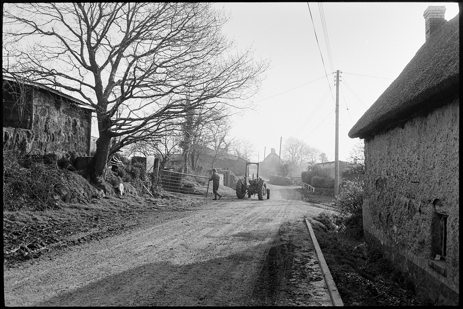 School bus arriving in village children waiting, morning. 
[A man closing a metal gate near a barn with hay bales and a road at Upcott, Dolton. A tractor is parked in the road and a thatched cob cottage can be seen in the foreground.]