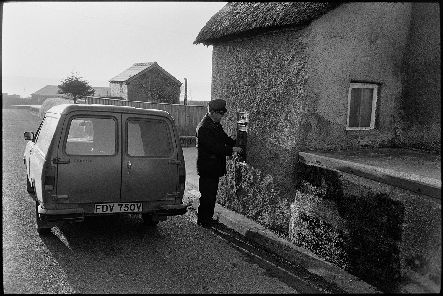 Postman emptying letterbox post, van parked, thatched cottage. 
[A postman collecting letters from a post box set into the wall of a cob cottage, which is also thatched, at Moor End, Burrington. His post van is parked in the road behind him.]