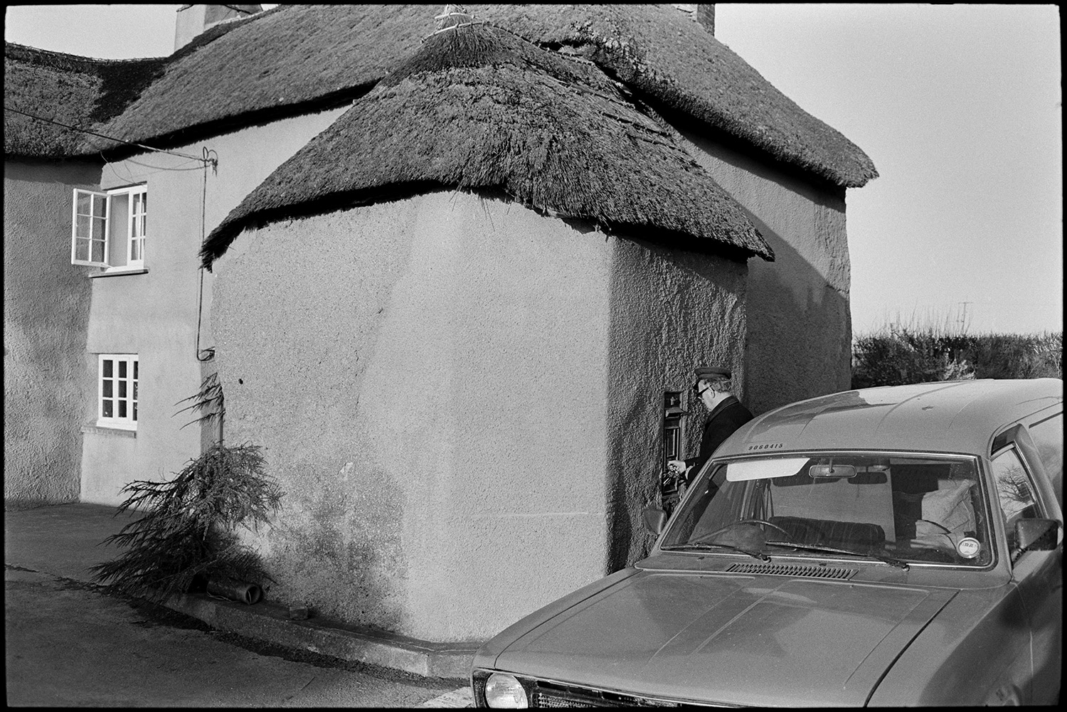 Postman emptying letterbox post, van parked, thatched cottage. 
[A postman locking a post box after collecting the letters inside, at Moor End, Burrington. The post box is set into the wall of a cob and thatch cottage. His post van is parked behind him on the road.]