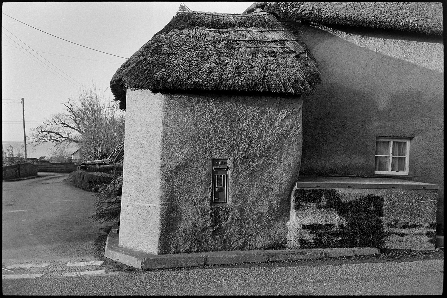 Postman emptying letterbox post, van parked, thatched cottage. 
[A post box set into the wall of a cob and thatch cottage at Moor End, Burrington.]