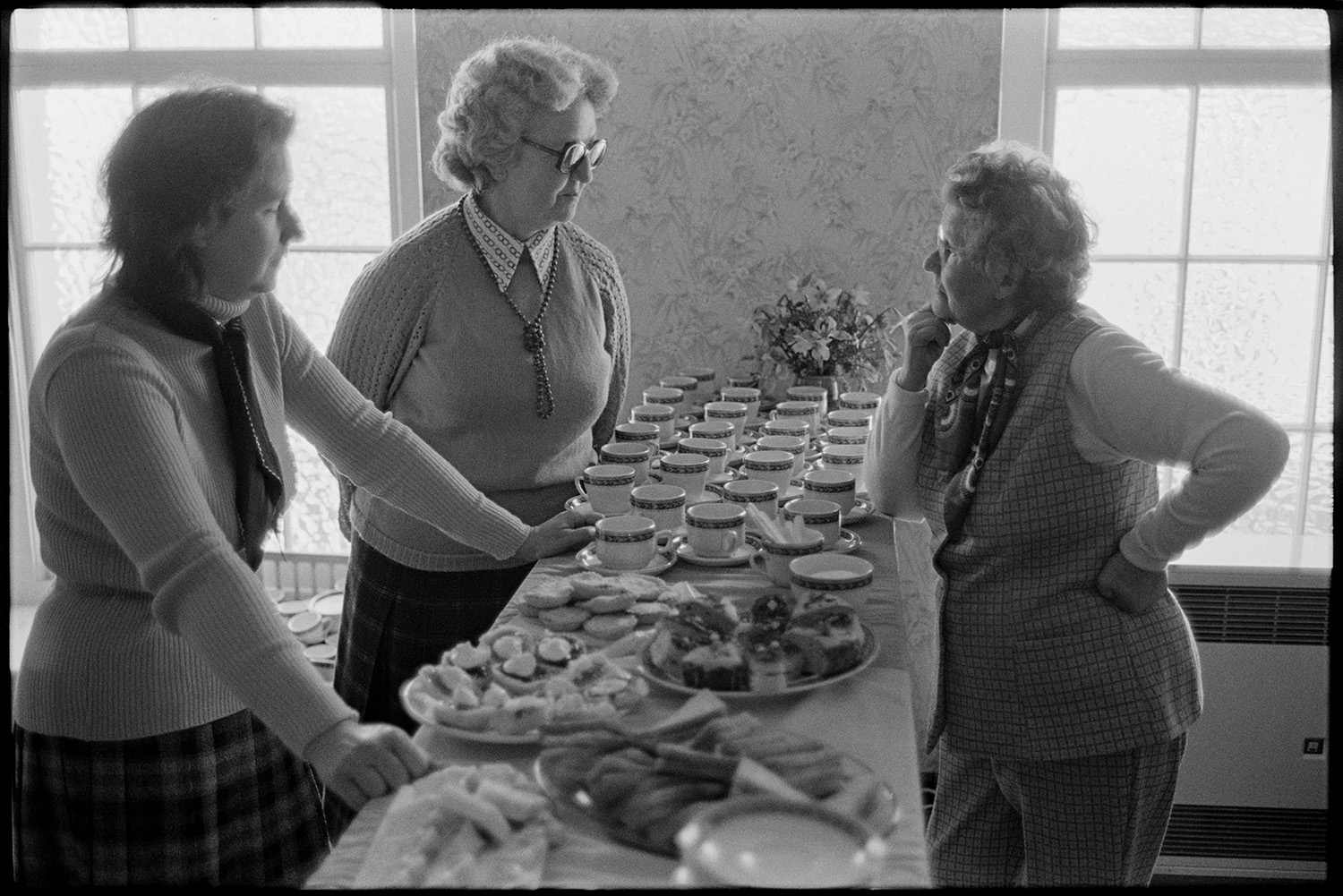 Women with tea and cakes at W I meeting, sandwiches. 
[Three women talking by a table laid with cakes, sandwiches and cups, at a Women's Institute meeting in the John Gay room at the Queen's Hall, Barnstaple.]