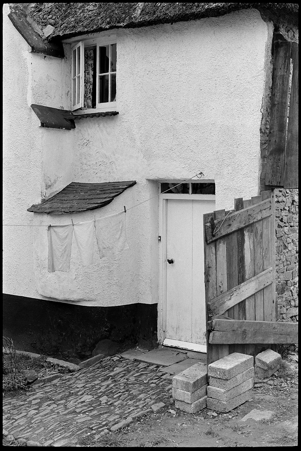Side of cottage now renovated and ruined, cobbles, bread oven. 
[The cobbled entrance to a thatched cottage in Beaford. A bread oven can be seen next to the front door and washing is hung out to dry on a washing line outside. The cottage was later renovated,]