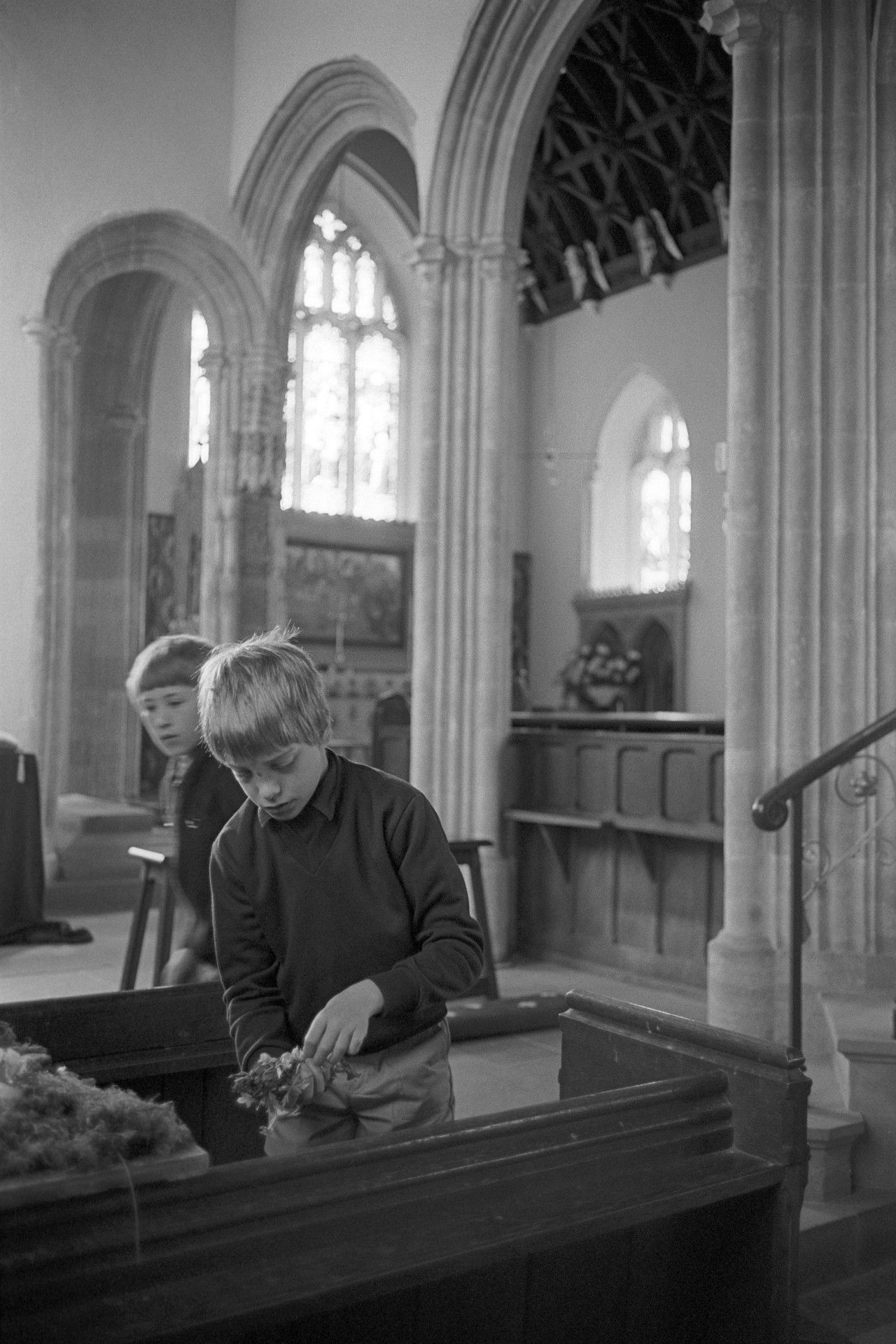 Boys arranging Church flowers. 
[Two boys arranging flowers in a pew in Chittlehampton Church. Arches and a side chapel can be seen in the background.]