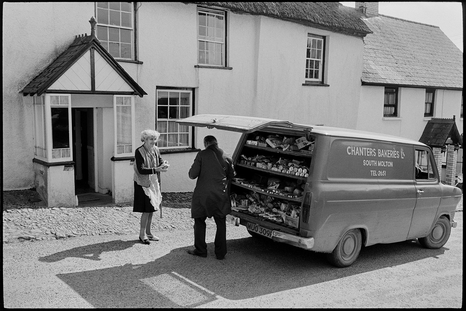 Baker selling from van. 
[A woman buying goods from a man with a Chanters Bakeries van, outside a cottage in Chittlehampton.]
