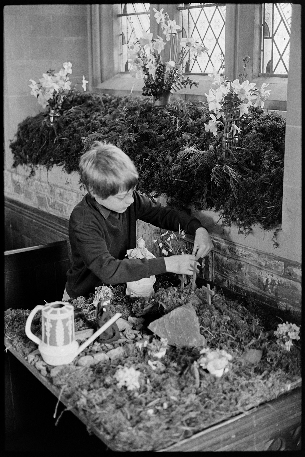 Boys arranging Church flowers. 
[A boy arranging flowers in an Easter Garden in Chittlehampton Church. A watering can is sat on the tray with the miniature garden. Another flower display can be seen in the windowsill behind him.]