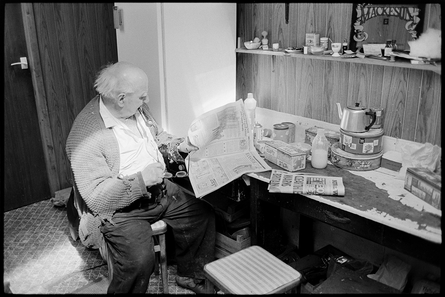 Man reading newspaper in his kitchen. 
[Archie Parkhouse reading a Sunday newspaper in his kitchen at West Lane, Dolton. He is smoking a pipe and holding a pair of glasses. Cake tins, a teapot and a bottle of milk are on the table in front of him.]