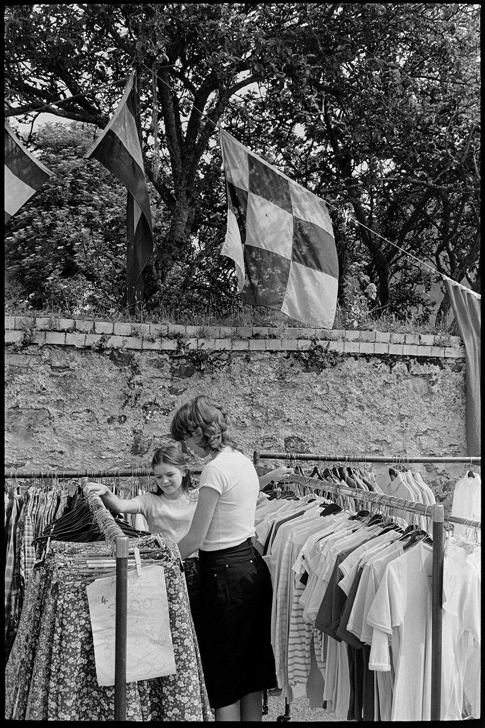 Open air market, village square part of festival, stalls, flags, crafts, books, plants. 
[A woman and child looking at a clothes stall at Dolton Festival or open air market. Flags are hung up from a tree behind a wall in the background.]