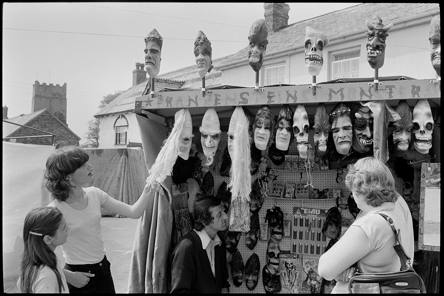 Open air market, village square part of festival, stalls, flags, crafts, books, plants. 
[Women and a child looking at masks on a stall called 'Frankenstein's Monster' at Dolton Festival or open air market. Dolton Church tower can be seen in the background.]