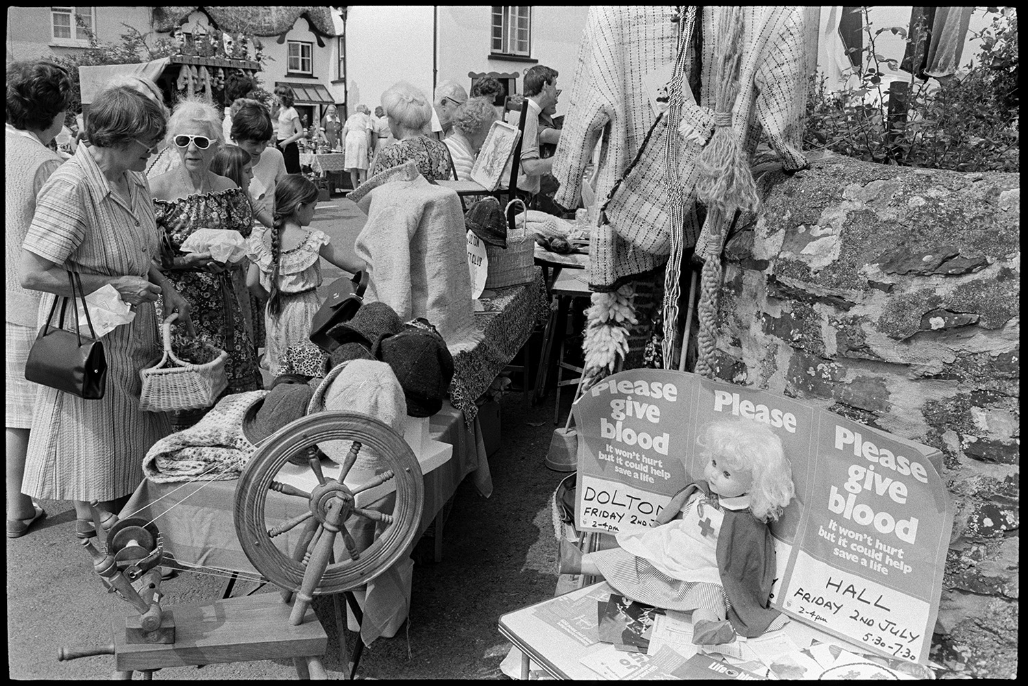 Open air market, village square part of festival, stalls, flags, crafts, books, plants. 
[Women and children looking at a stall with clothes and hats at Dolton Festival or open air market. A spinning wheel is next to stall along with posters asking people to give blood.]