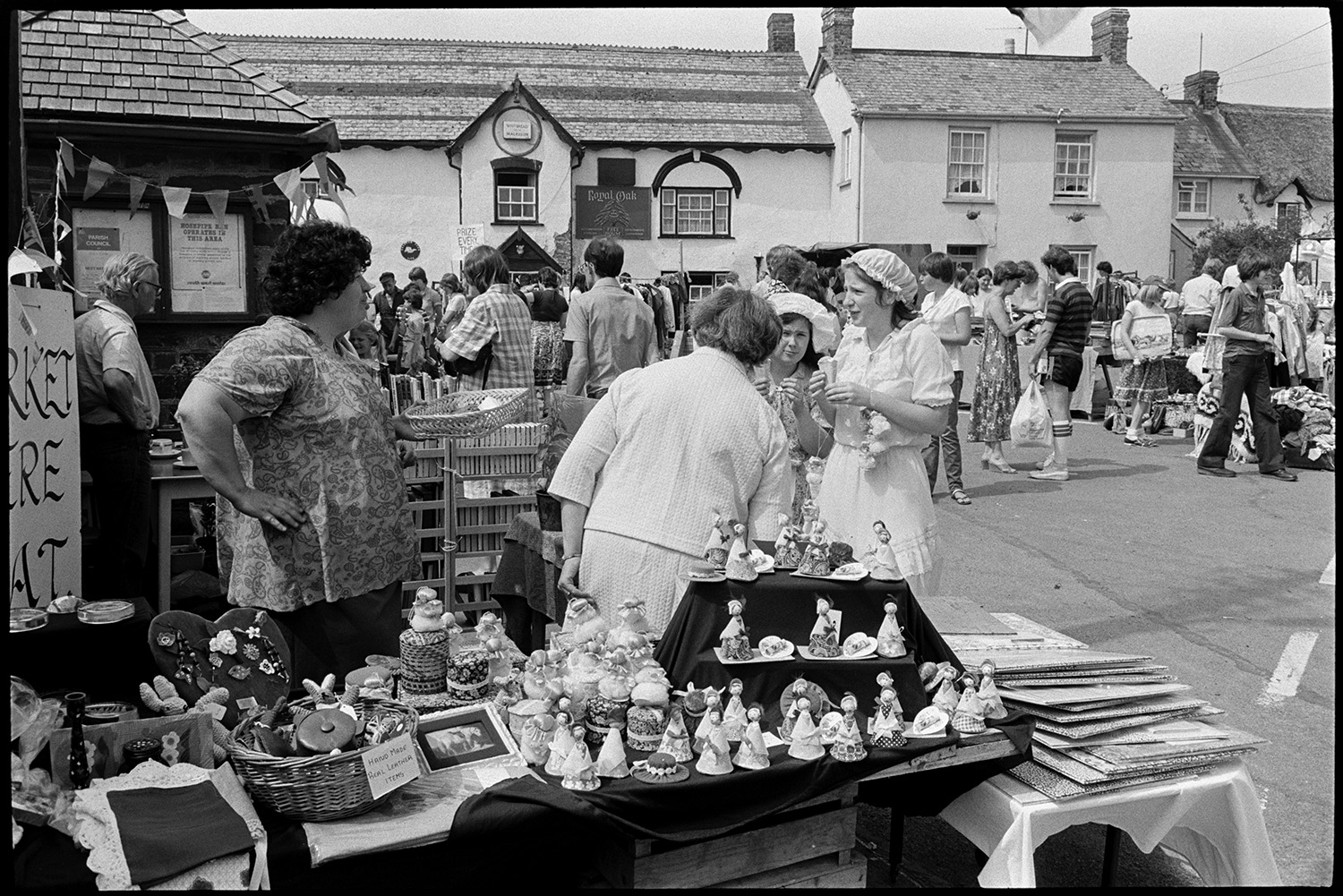 Open air market, village square part of festival, stalls, flags, crafts, books, plants. 
[A woman running a stall selling broaches, knitted items and small figurines at Dolton Festival or open air market outside the Royal oak pub in Dolton. People are looking at other stalls in the background.]