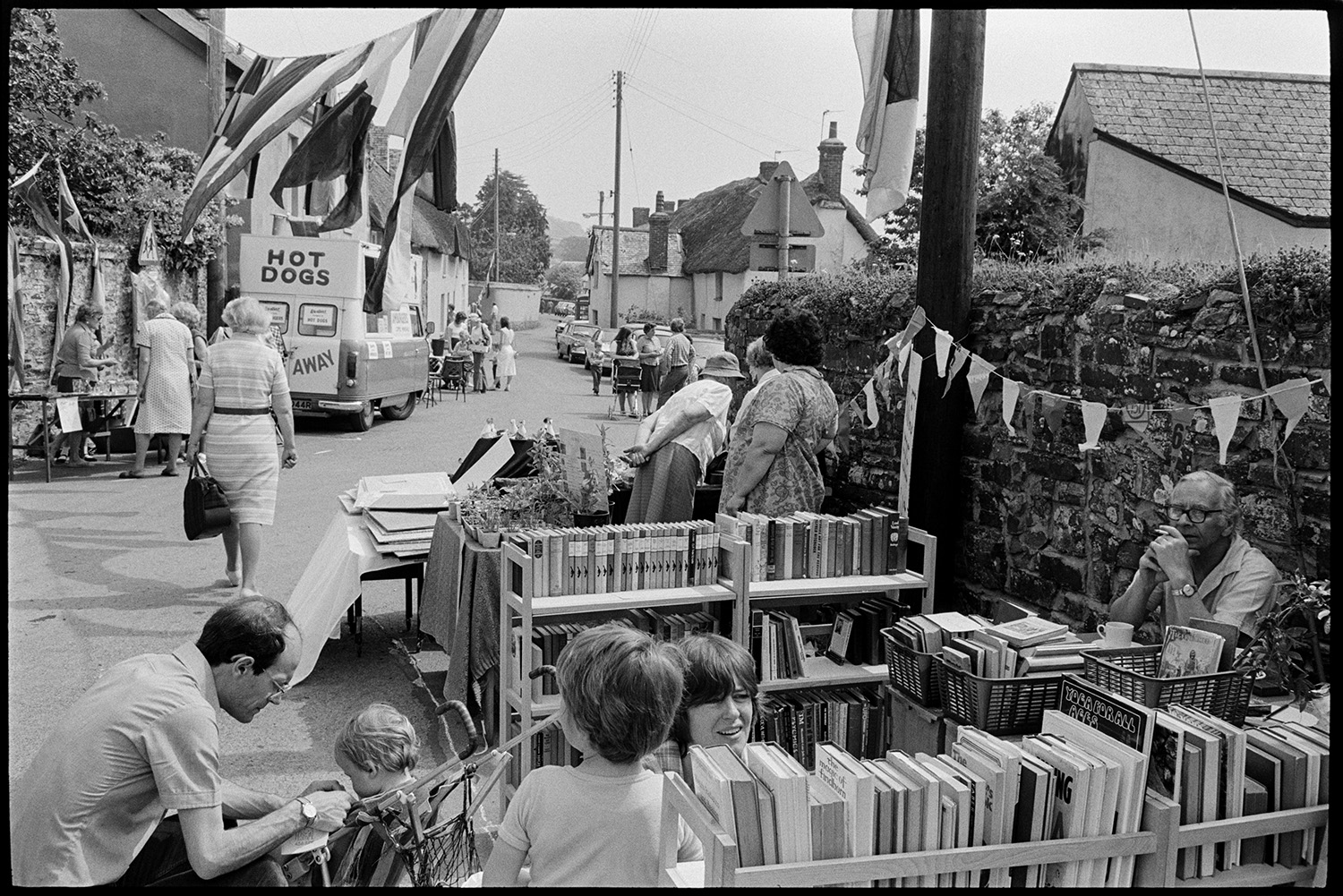 Open air market, village square part of festival, stalls, flags, crafts, books, plants. 
[A family looking at a book stall at Dolton Festival or open air market. Bunting is hung up behind the stall and flags are suspended between lamp posts. A hot dog van can be seen in the background.]