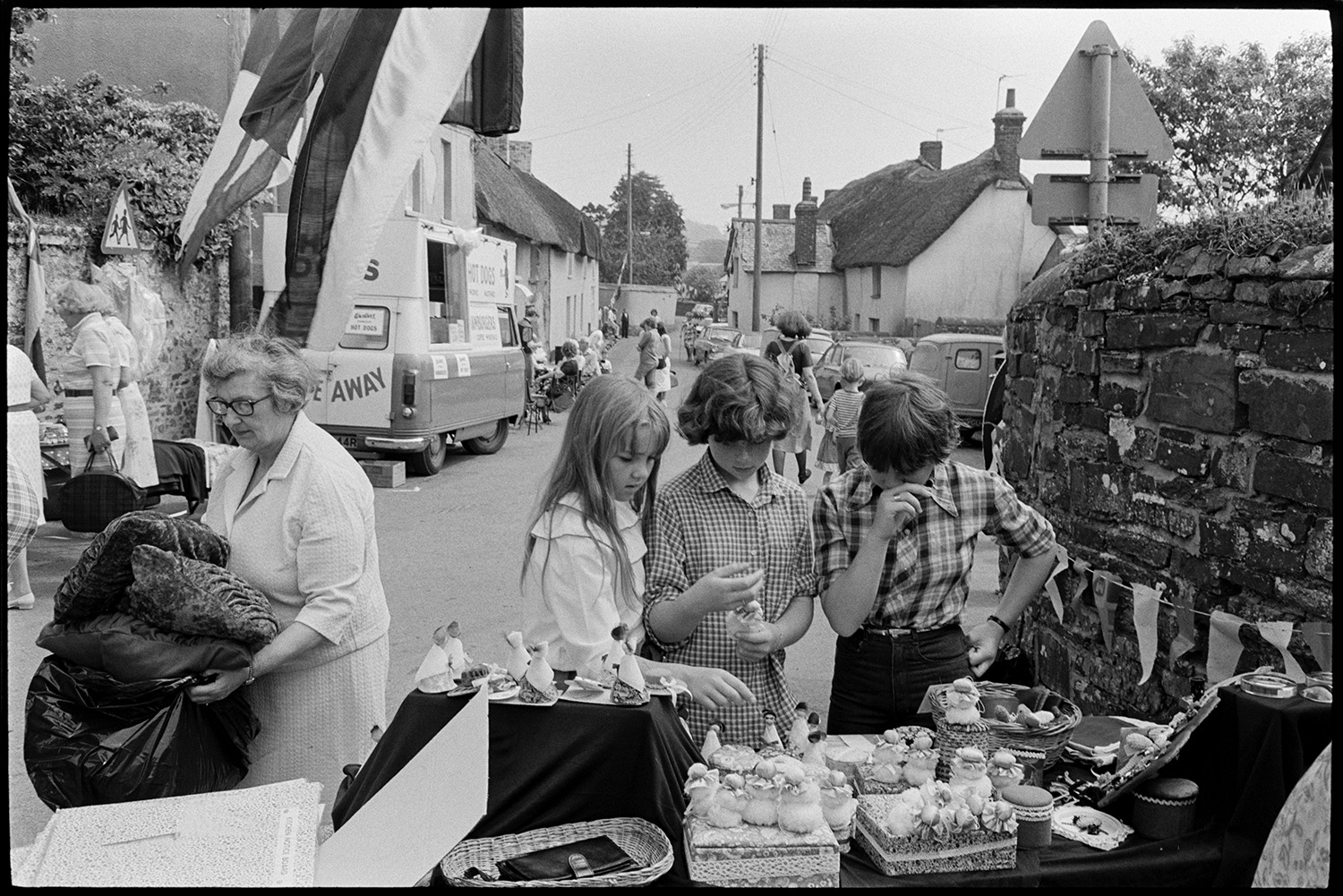 Open air market, village square part of festival, stalls, flags, crafts, books, plants. 
[Three children looking at a stall selling knitted items and small figurines at Dolton Festival or open air market. A woman is walking past with a bin bag full of cushions. The street is decorated with bunting and flags, and a hot dog van can be seen in the background.]