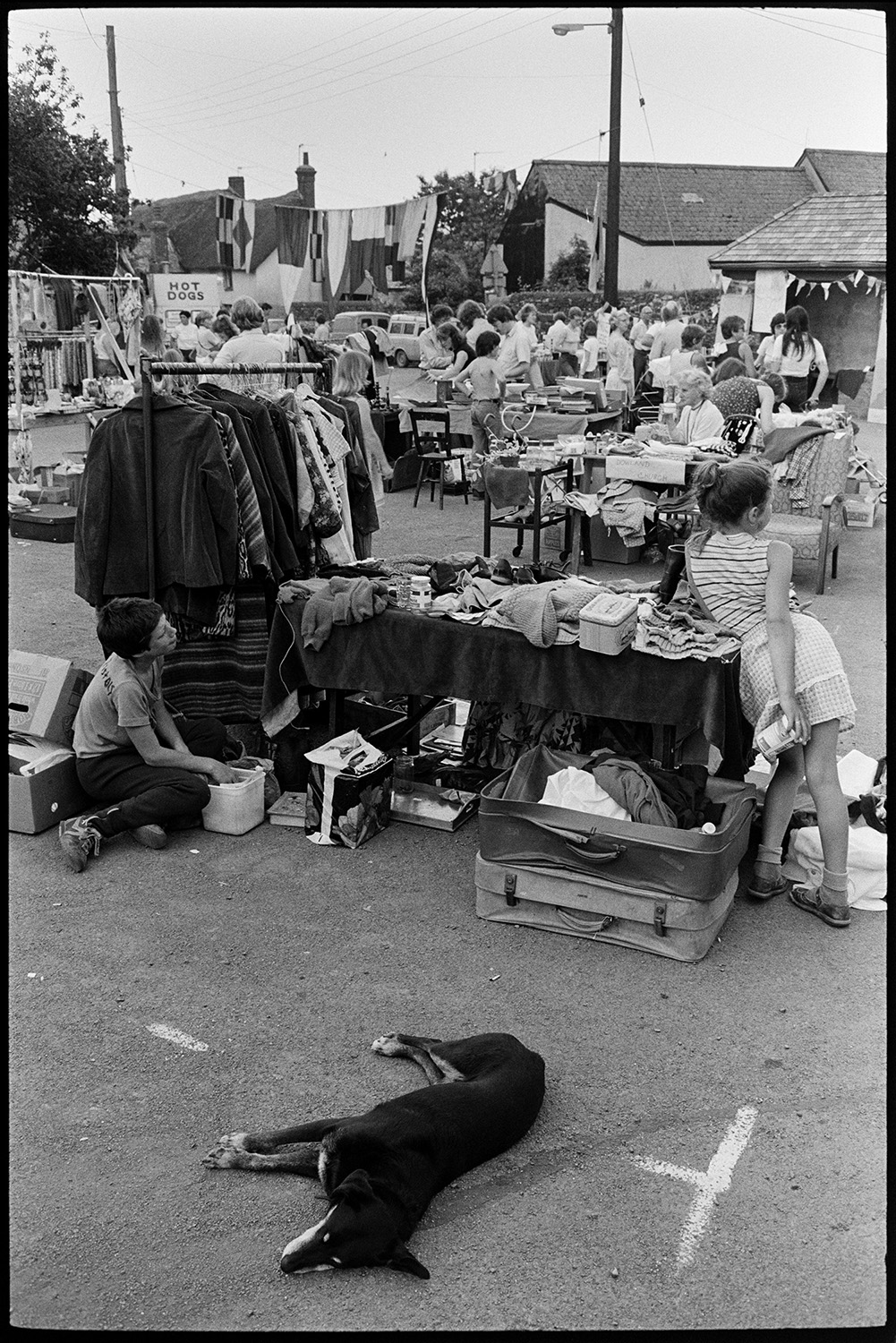 Open air market, village square part of festival, stalls, flags, crafts. 
[A dog lying on the ground by a stall selling clothes at Dolton Festival or open air market. Two children are running the stall. Other stalls are visible in the background and the street is decorated with flags.]