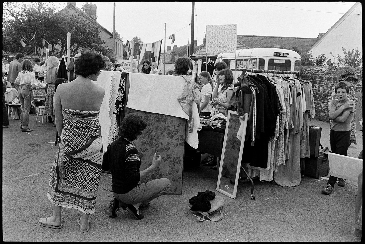 Open air market, village square part of festival, stalls, flags, crafts. 
[Two people looking at themselves in a mirror by a clothes stall at Dolton Festival or open air market. Other stalls can be seen in the background and the street is decorated with flags.]