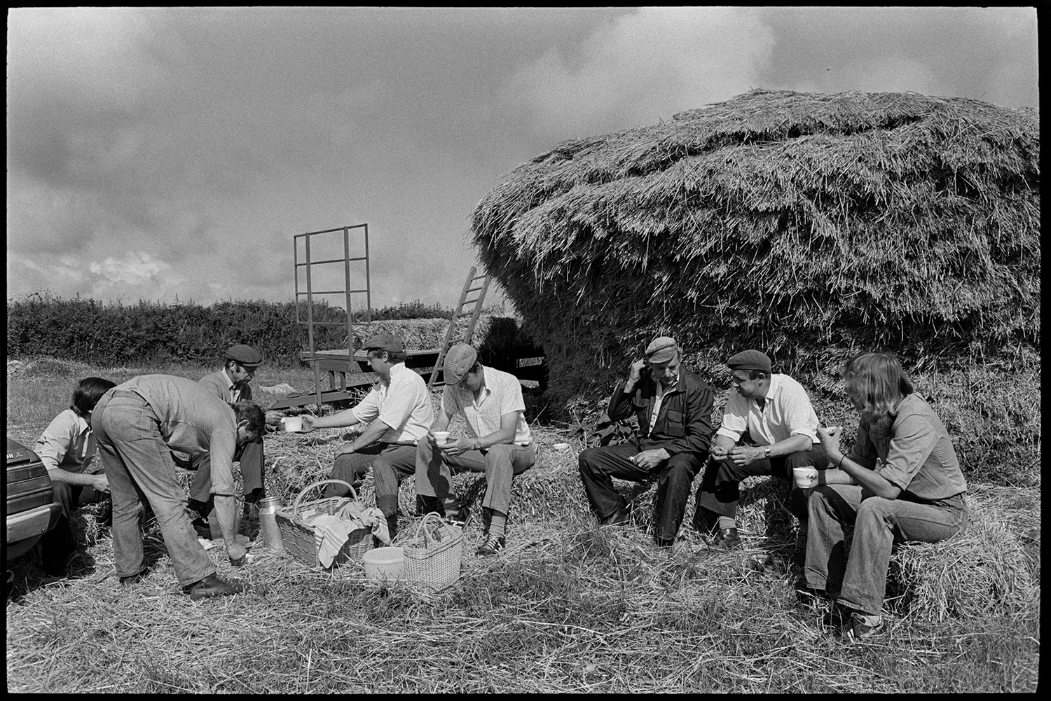 Reedcombing machine, nitches of reed, men working in sunlight. 
[Stephen Middleton and other men having a tea break from reedcombing, in a field at Westacott, Riddlecombe. They are sat on hay bales in front of the rick. Baskets of food and a flask of tea are on the ground in front of them. A trailer and ladder can be seen in the background.]