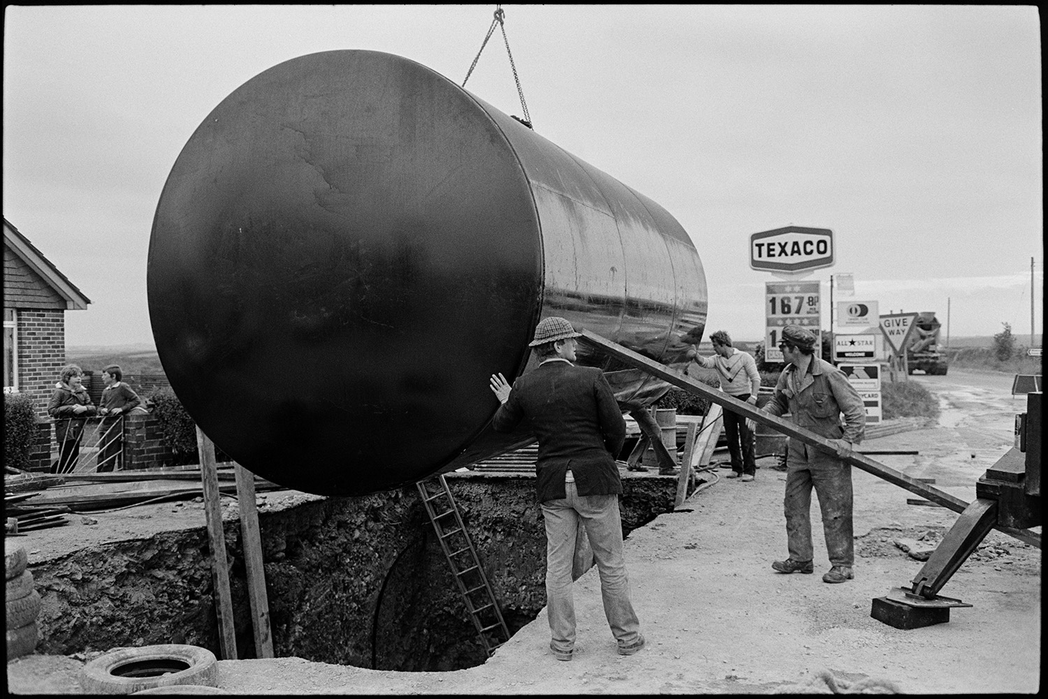 Men with crane installing huge underground petrol tank at garage.
[Four men supervising the lowering of a large petrol tank by crane into an excavated hole at Beacon Garage, Dolton Beacon. Two boys are watching from gate to an adjoining building, and a Texaco petrol sign is visible in the background.]