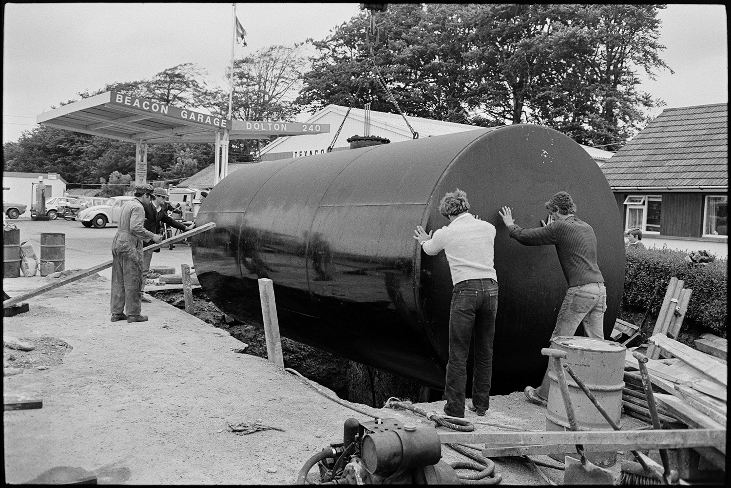 Men with crane installing huge underground petrol tank at garage.
[Four men helping to lower a large petrol tank being lowered by a crane into an excavated hole at Beacon Garage, Dolton Beacon. Cars on the forecourt of the Garage are visible in the background.]