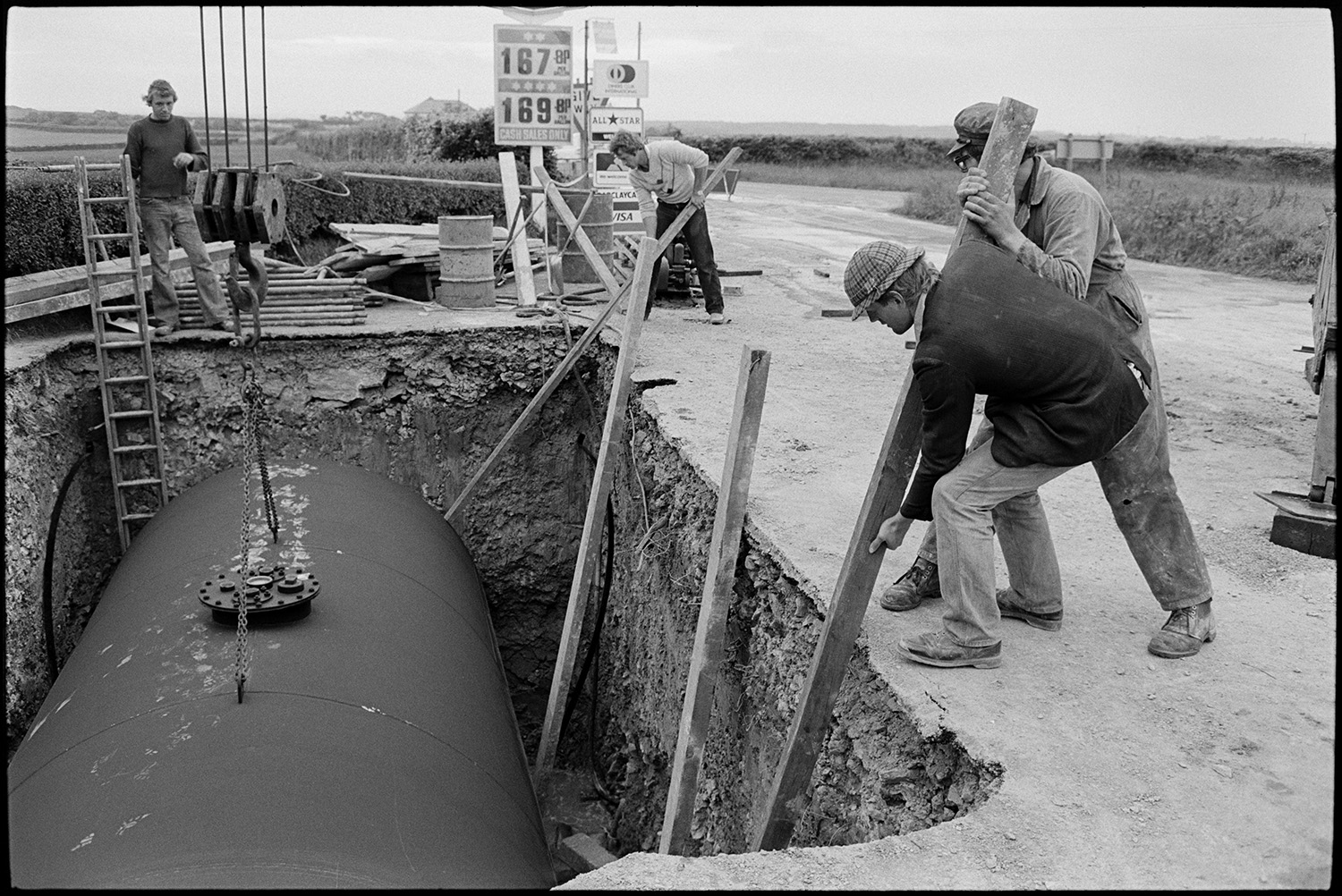 Men with crane installing huge underground petrol tank at garage.
[Four men installing a large petrol tank into an excavated hole at Beacon Garage, Dolton Beacon. They are using long wooden poles as levers.]