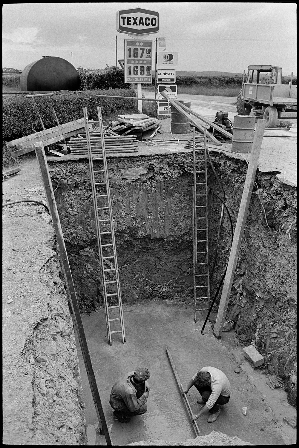 Men with crane installing huge underground petrol tank at garage.
[Two men at Beacon Garage, Dolton Beacon working in a deep excavated hole fitting an underground petrol tank. A lorry and a Texaco garage sign are visible in the background.]