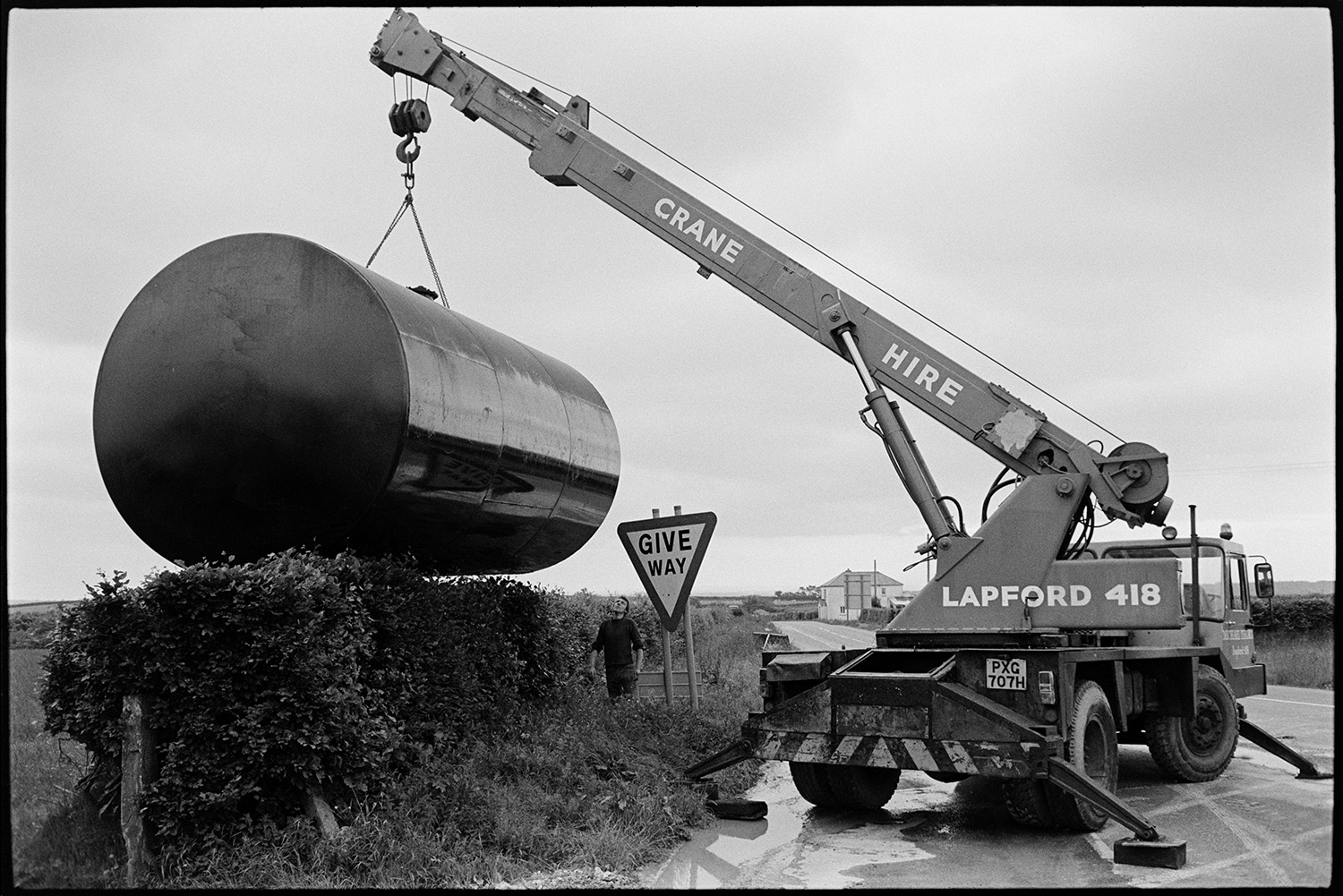 Men with crane installing huge underground petrol tank at garage.
[A man at Beacon Garage, Dolton Beacon standing next to a Give Way sign supervising the movement of a large petrol tank over a hedge next to the road, using a crane.]