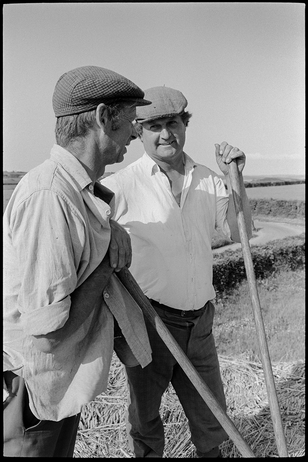 Two reedcombers resting and chatting, wearing caps.
[Two men reedcoming. They are wearing caps and leaning on their pitchforks chatting in the bright sunshine at Westacott, Riddlecombe.]