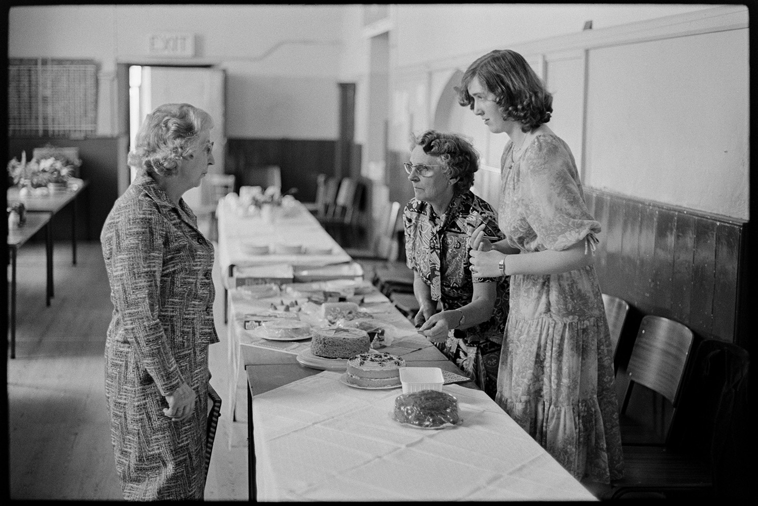Stalls in village hall, women setting out cakes and produce.
[Three women laying out cakes on tables covered with tablecloths in Roborough Village Hall for Roborough Fair.]