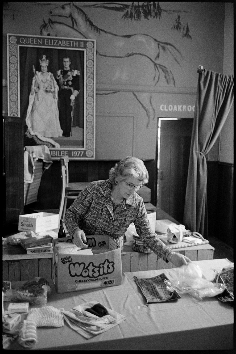 Stalls in village hall, women setting out cakes and produce.
[A woman setting out products on a cloth covered table in Roborough Village Hall, for Roborough Fair. A wall mural of horses is visible on the wall in the background, behind a large picture of Queen Elizabeth II and the Duke of Edinburgh commemorating her Silver Jubilee in 1977.]