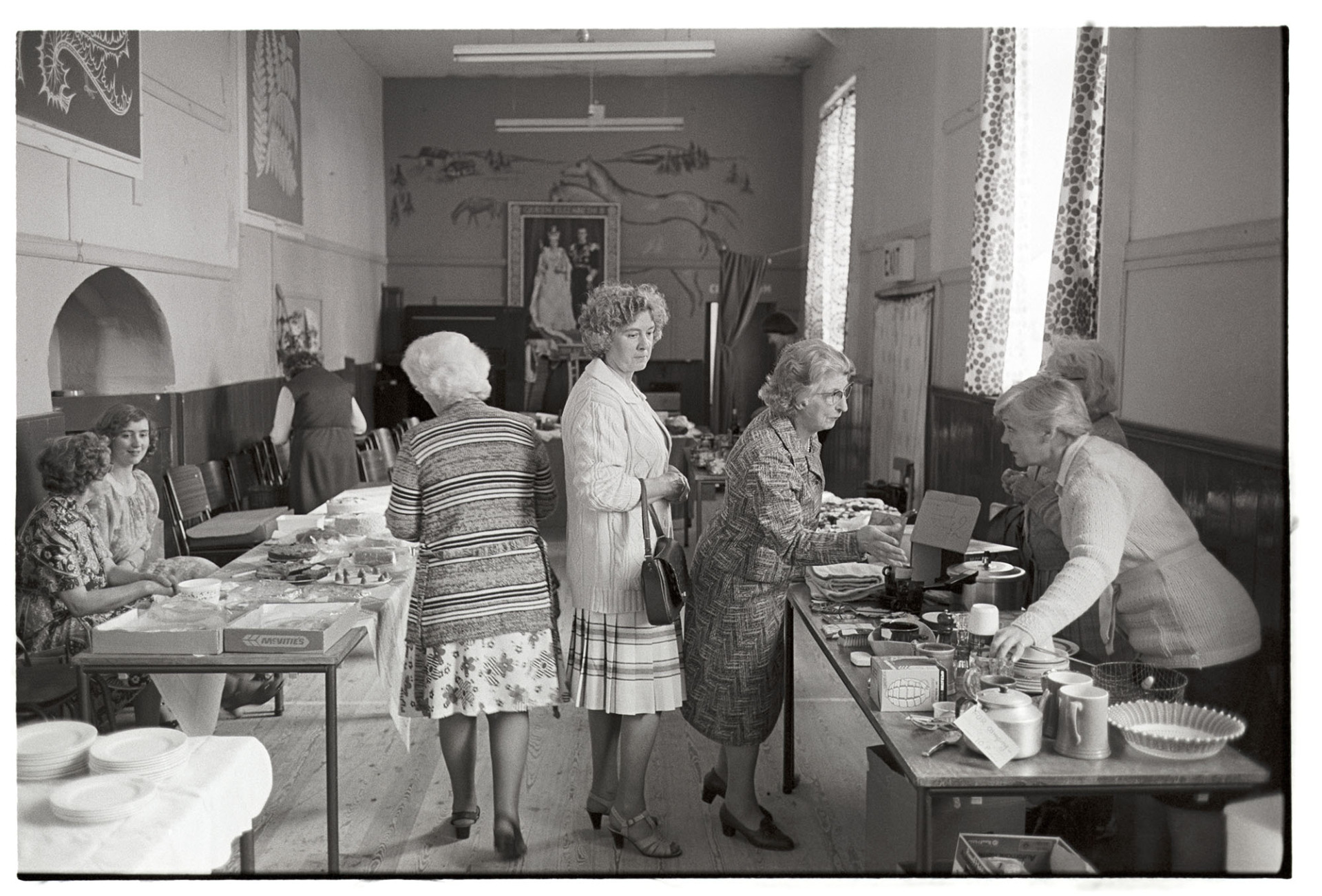 Stalls in village hall, women looking at cakes and produce. Dresses and cardigans. 
[Women looking at cake and bric-a-brac stalls at Roborough Fair in Roborough Village Hall. The walls are decorated with pictures, including a royal portrait at the far end of the hall.]