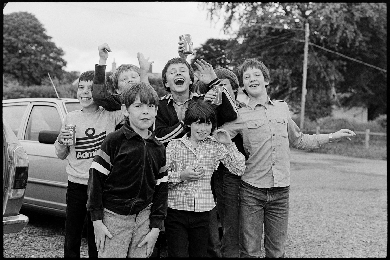Crowd of children laughing.
[A group of children in a car park, holding cans of Coke, laughing and waving to the camera.]