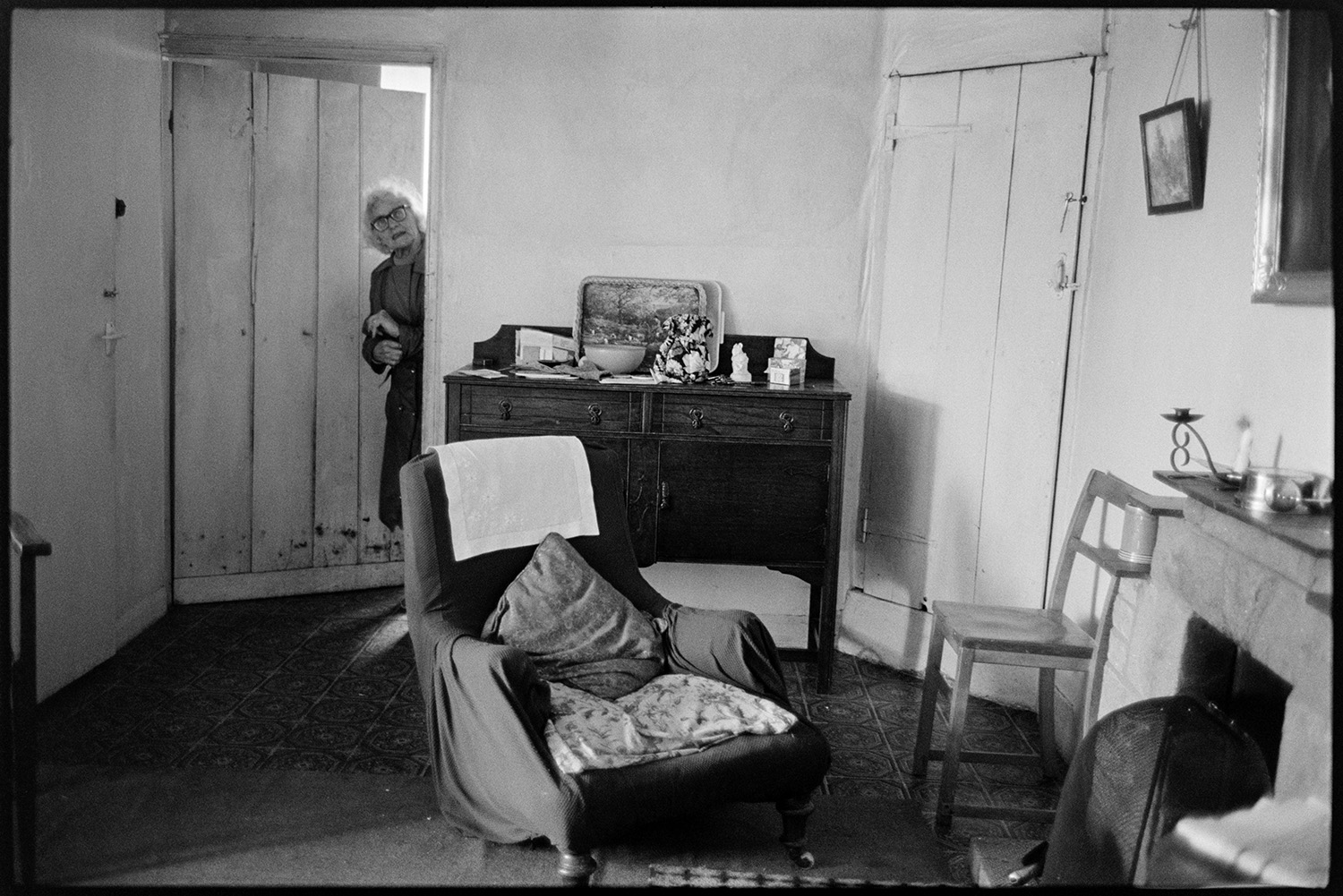 Women chatting and making tea in room with piano, television. 
[A woman looking through a doorway into a room in a house in Roborough. Various items of furniture can be seen including a chair, fireplace and sideboard.]