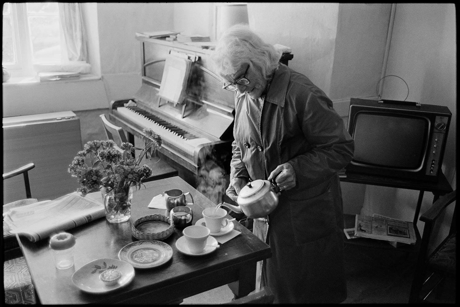 Women chatting and making tea in room with piano, television. 
[A woman pouring cups of tea in a house in Roborough. A piano an television can be seen in the room and a vase of Sweet William flowers are on the table.]