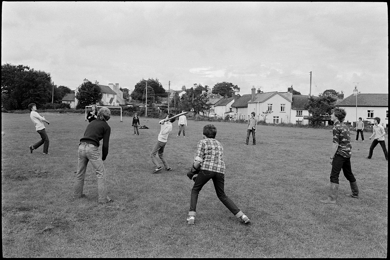 Young people playing cricket on village playing field. 
[A group of teenage boys playing cricket on the village playing field at Dolton. Houses can be seen in the background.]